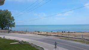Una carretera costera con una playa a la derecha, que muestra una calzada pavimentada, una zona cubierta de césped y una playa arenosa con rocas y una valla.