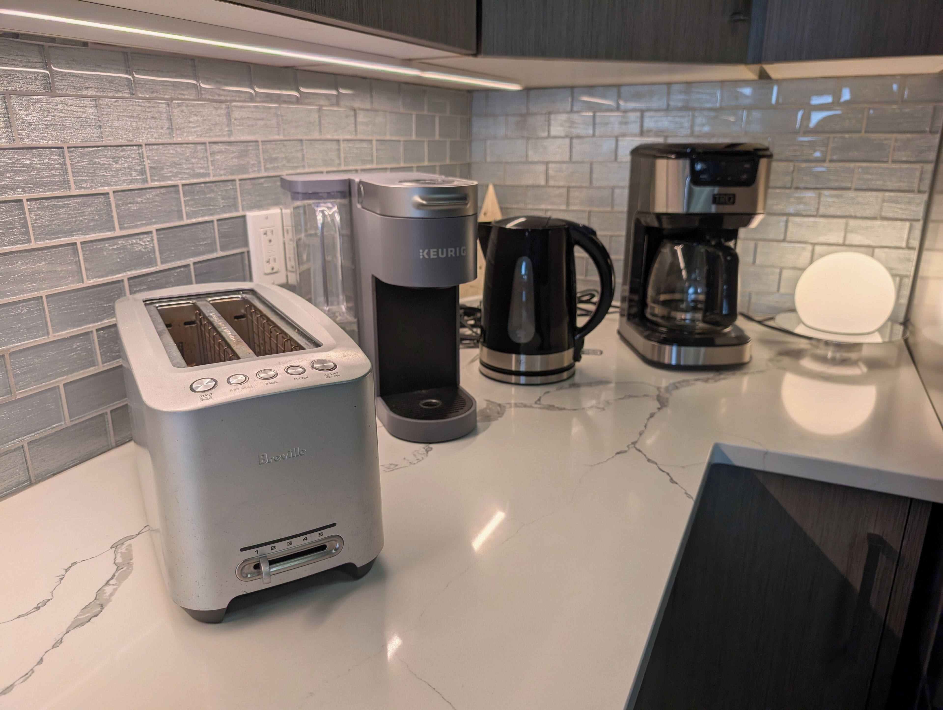 A kitchen with a Breville toaster, a Keurig coffee maker, and a black coffee maker on a marble countertop.
