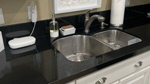 Una cocina con encimera negra, armarios blancos, pileta de acero inoxidable y un panel decorativo sobre la pileta.