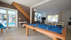 Et billardbord i et rum med trappe, spiseområde og en swimmingpool, der ses gennem glasdøre.