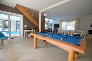 A pool table in a room with a staircase, a dining area, and a pool visible through glass doors. - Villa with indoor pool, billiards, sauna, 230m2, Seafront, Jard sur Mer (Jard-sur-Mer)