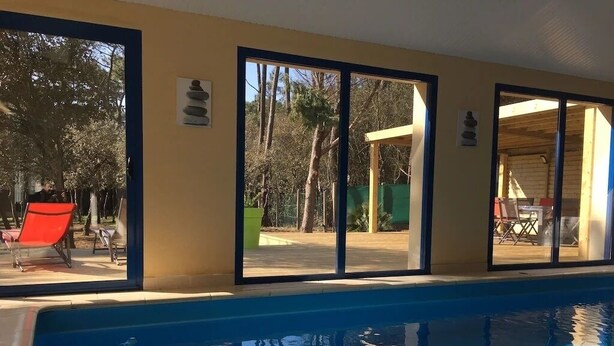 En swimmingpool med glasvægge og -døre, en rød stol og udsigt til træer og en bygning.
