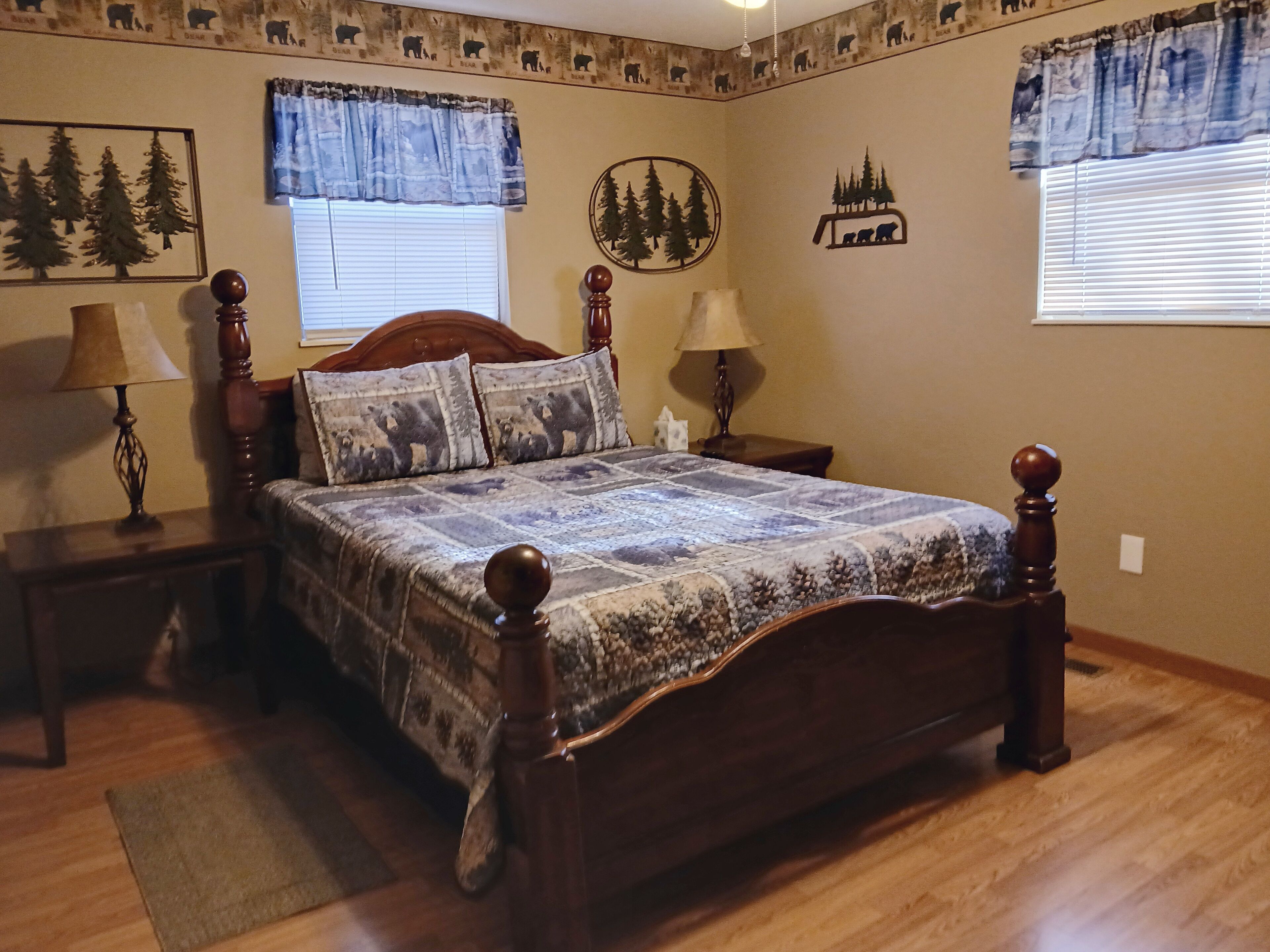 A bedroom with a wooden bed, two bedside tables, a nightstand, a rug, and wall decorations including a bear-themed picture and a forest mural.