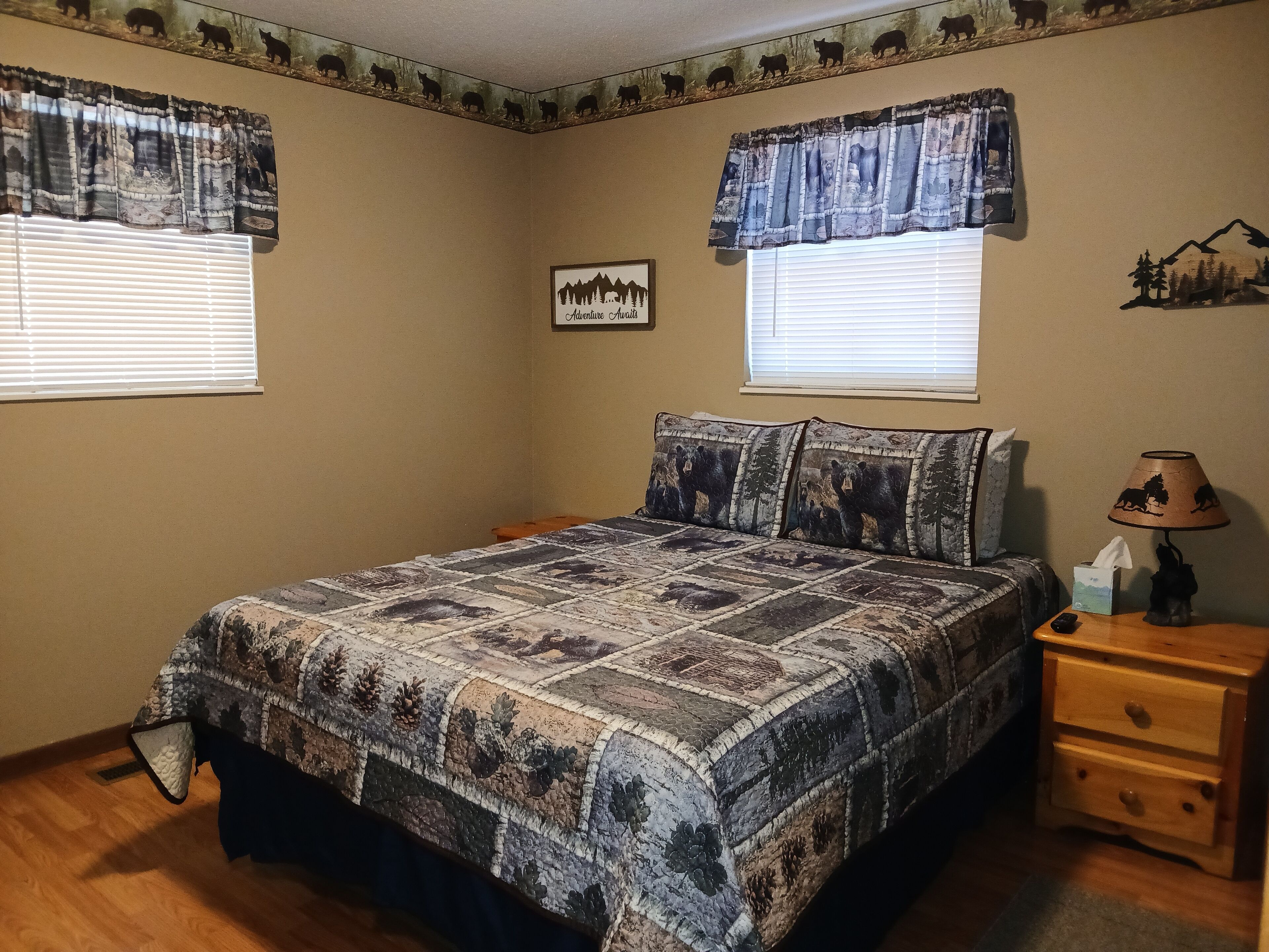 A bedroom with a large bed, two windows with curtains, a bedside table with a lamp, and a wall-mounted picture.