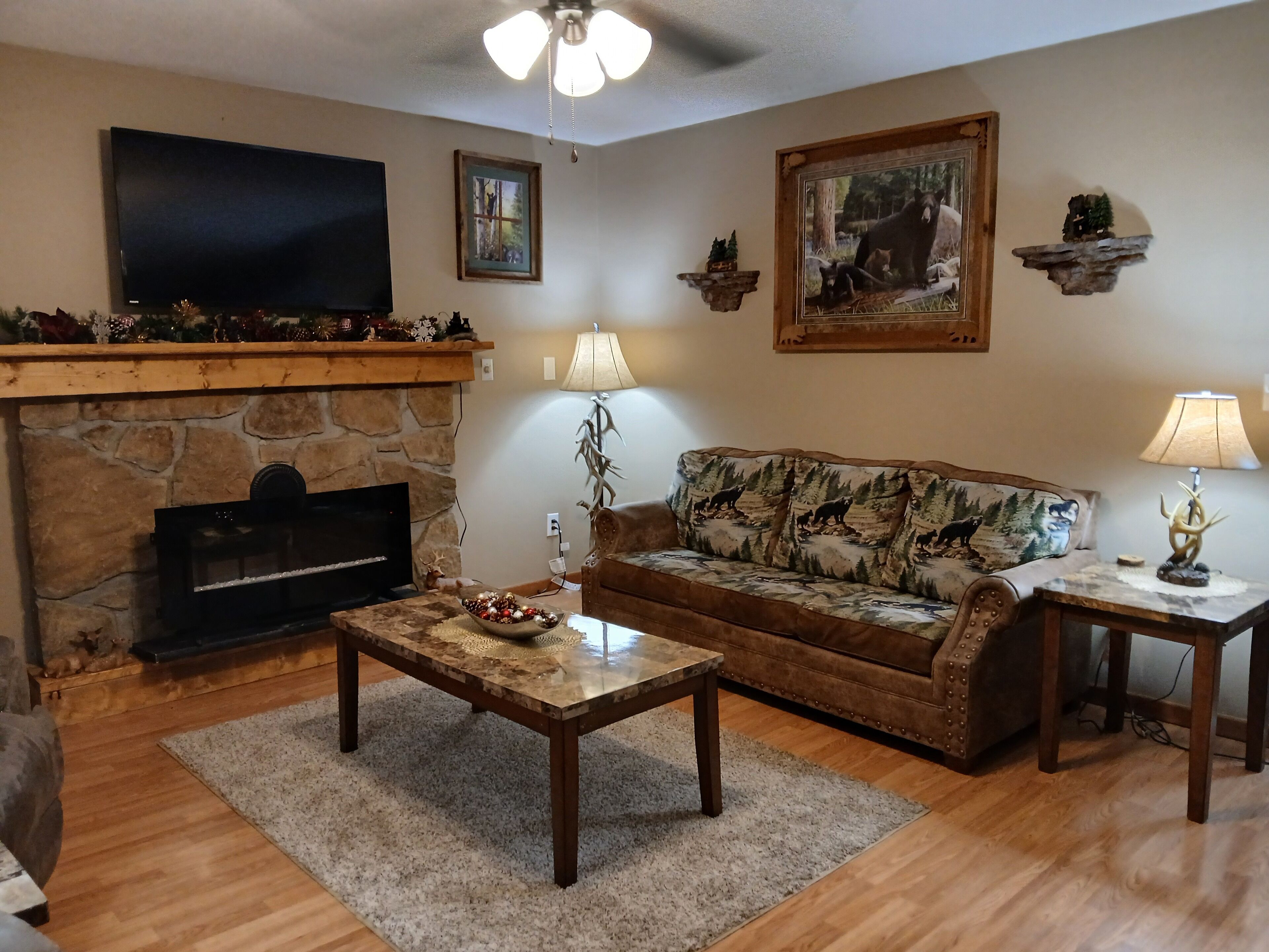 A living room with a stone fireplace, a flat-screen TV, a couch with a forest print, and two lamps.