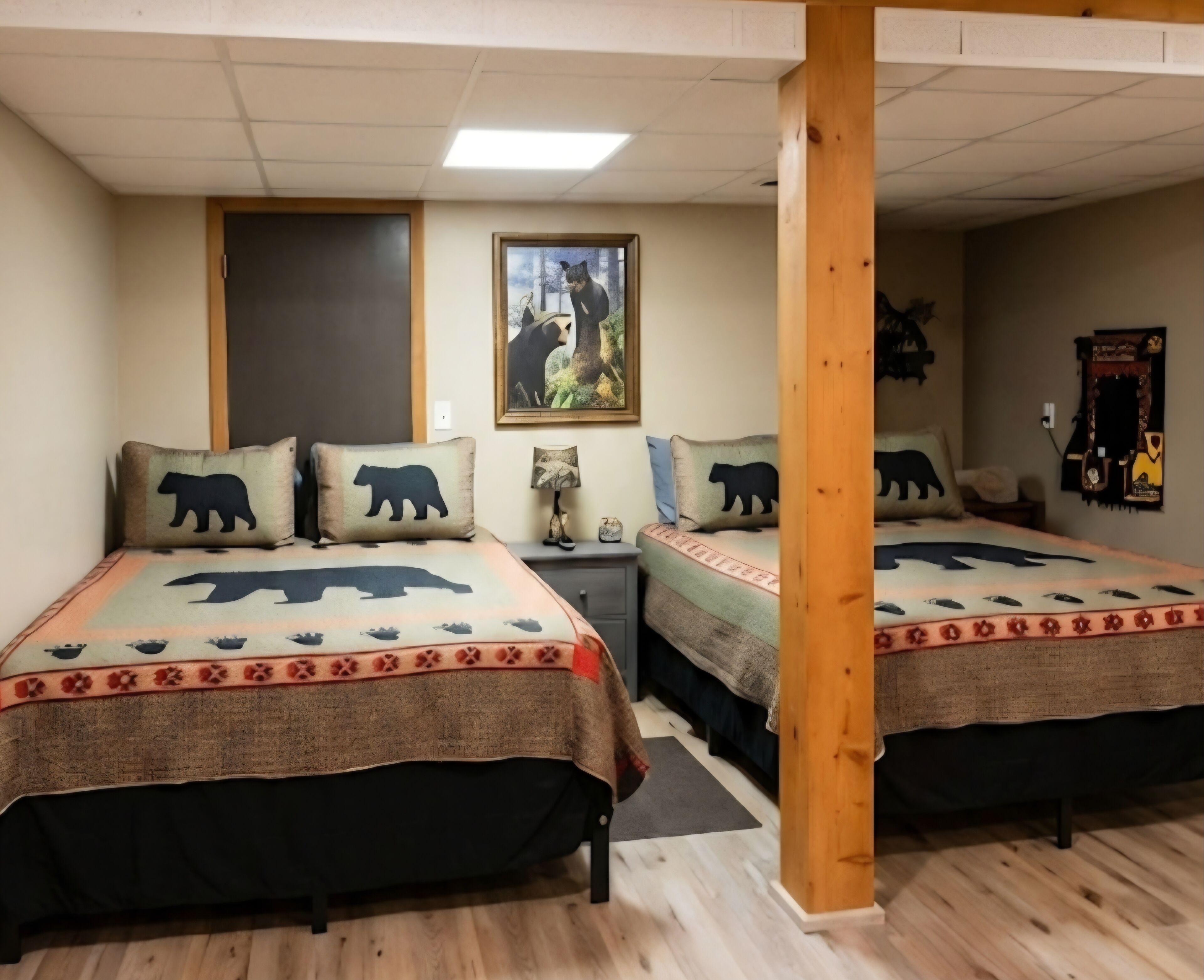 A room with two beds, each with a bear-themed bedspread and matching pillows. There is a wooden column in the center, a painting on the wall, and a small table with a lamp.