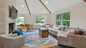 A spacious living room with a stone fireplace, large windows, a cozy sofa, and a rustic wooden chest. - Spacious 4BR/3BA with bunkhouse; perfect for family and friends! (Bryan)
