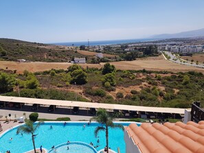 Ein Poolbereich mit Schwimmern und spielenden Personen, umgeben von hügeliger Landschaft und Gebäuden in der Ferne.