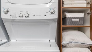 A front-loading washing machine with a control panel, a wooden shelf unit with folded towels, a pillow, and a basket.