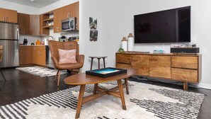 A modern kitchen with wooden cabinets and stainless steel appliances, a dining area with a wooden table and chairs, a flat-screen TV, and a small side table.
