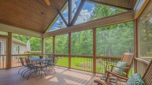 A covered outdoor deck with a wooden table and chairs, a wooden bench with cushions, and a view of a green landscape through large windows.