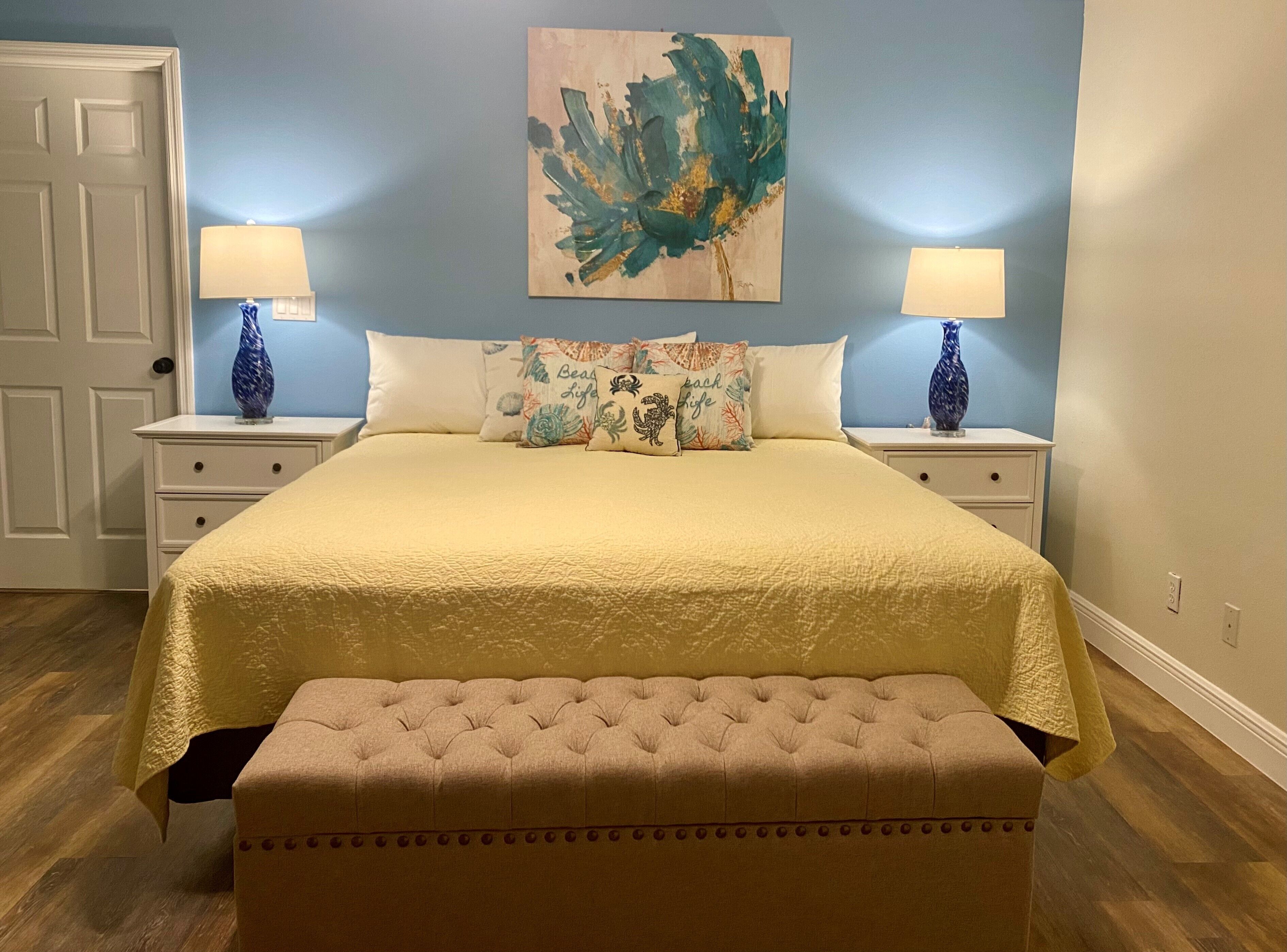 A bedroom with a large bed, two nightstands with lamps, a bench, and a wall painting.
