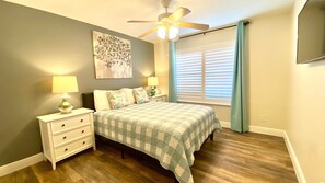 Quarto com cama, mesas de cabeceira, ventilador de teto, janela com persianas e uma obra de arte na parede.