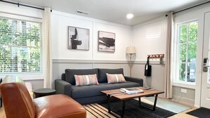 A modern living room with a grey sofa, a brown leather chair, and a coffee table. There are two framed artworks on the wall, a floor lamp, and a window with blinds.