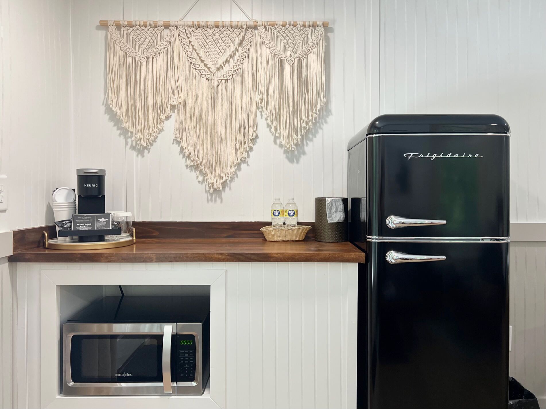 A kitchen with a black Frigidaire refrigerator, a microwave, a coffee maker, and a macrame wall hanging.