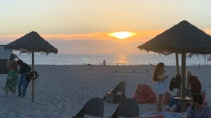 Cena de praia com guarda-sóis de palha, cadeiras de praia e pessoas relaxando.
