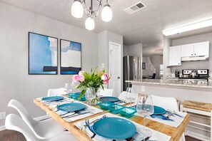 Uma sala de jantar moderna com uma mesa de madeira posta para seis pessoas, cadeiras brancas e uma cozinha ao fundo.