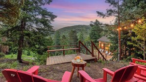 A wooden deck with red chairs and a table, overlooking a forested area with a small cabin in the background. -     Private Luxury Retreat in Evergreen, Hot Tub + Multiple Decks (Evergreen)