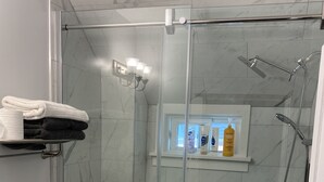Moderni kylpyhuone, jossa on lasiseinällä varustettu suihku, wc ja hylly, jossa on hygieniatuotteita.