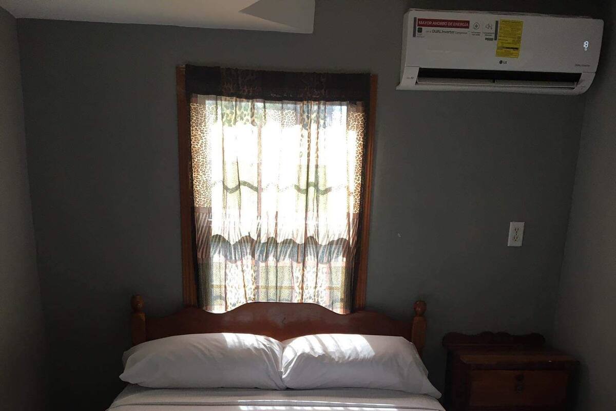 A bedroom with a bed, a nightstand, a window with patterned curtains, and an air conditioning unit.