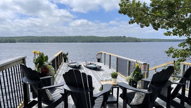 Eine Holzterrasse mit schwarzen Adirondack-Stühlen und einem Tisch mit Blick auf einen See, auf dem Boote fahren, und einem bewaldeten Ufer.