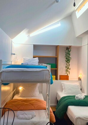 A dormitory room with bunk beds, a bedside table with a lamp, a shelf with books, and a hanging plant. - Naval Guest House & Bistrô (Vila do Conde)