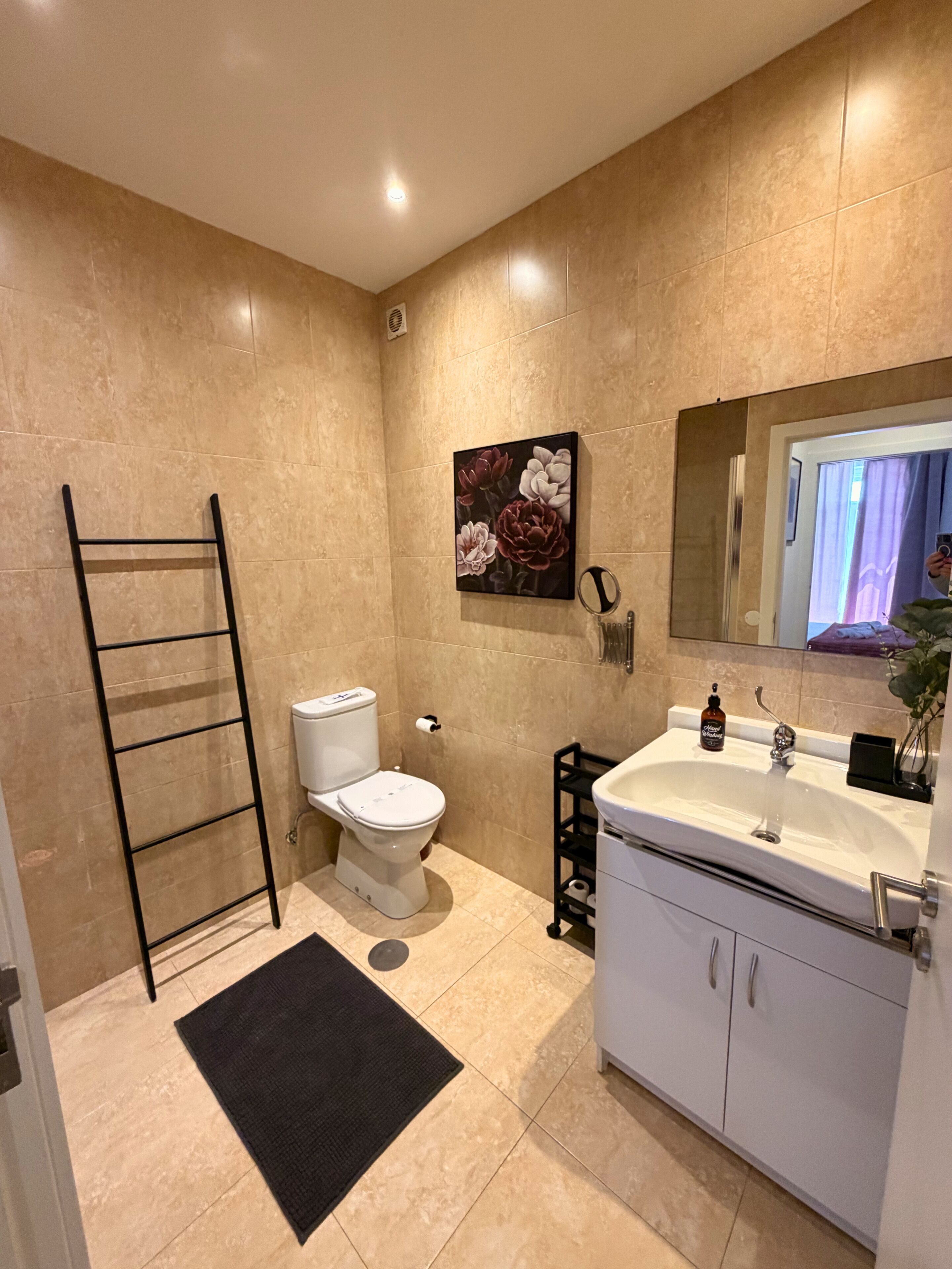 A bathroom with beige tiles, a white toilet, a black towel rack, a sink with a mirror, and a floral painting on the wall.