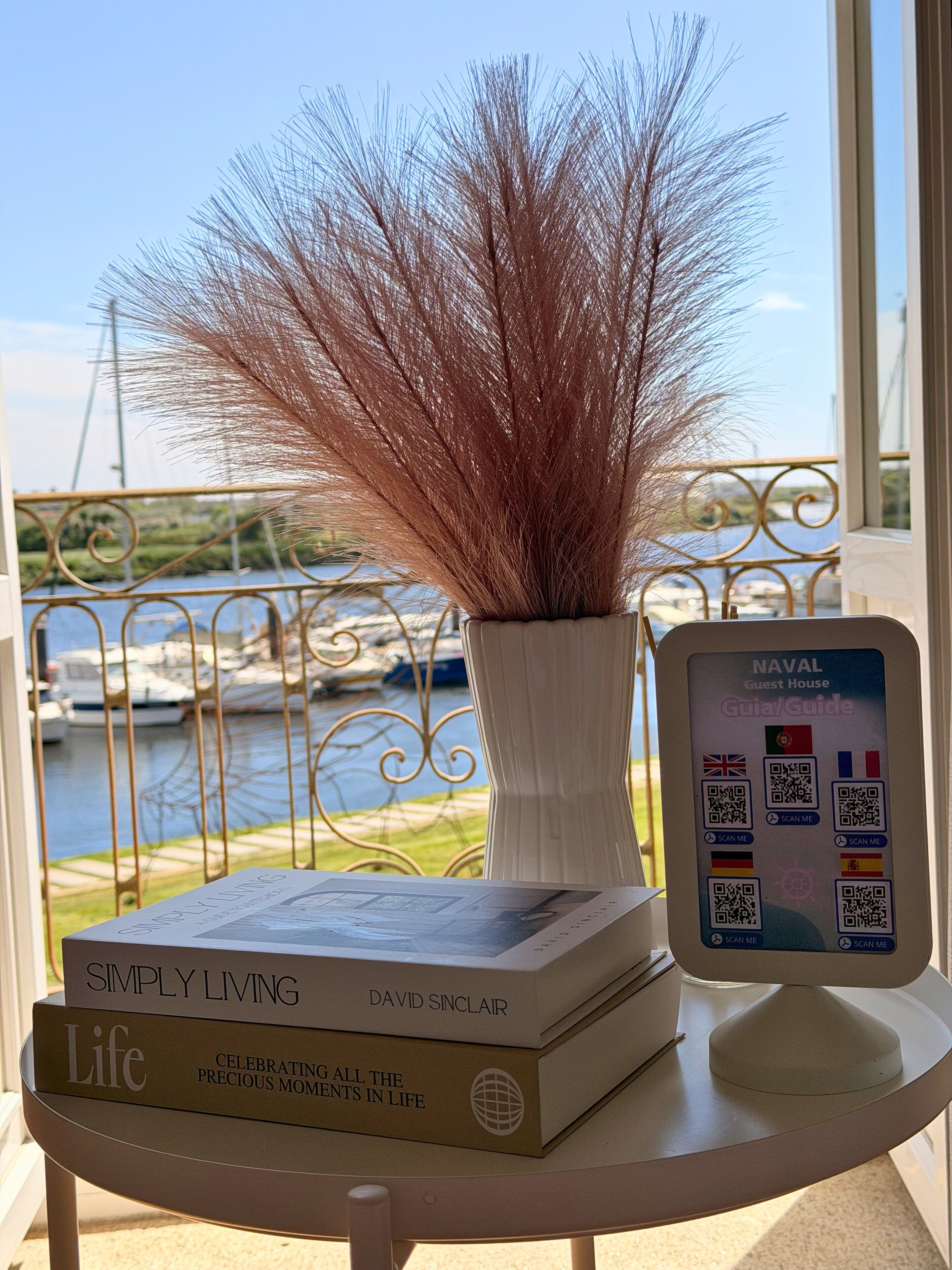 A table with a book titled 'Simply Living' by David Sinclair, a potted plant, and a QR code sign for the 'NAVAL Guest House.'