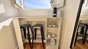 Una kitchenette compacta con dos taburetes, una estantería con utensilios de cocina y una ventana arriba.