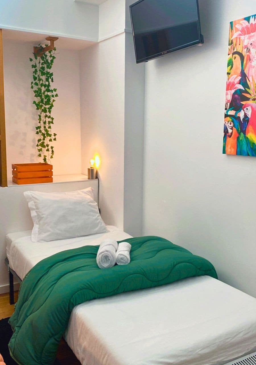 Una habitación pequeña y ordenada con una cama, una mesita de noche con lámpara, un televisor montado en la pared y un cuadro de colores.