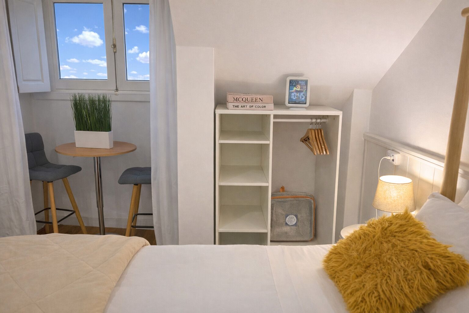 A bedroom with a bed, a bedside lamp, a small table with a plant, a wardrobe, and a window with curtains.