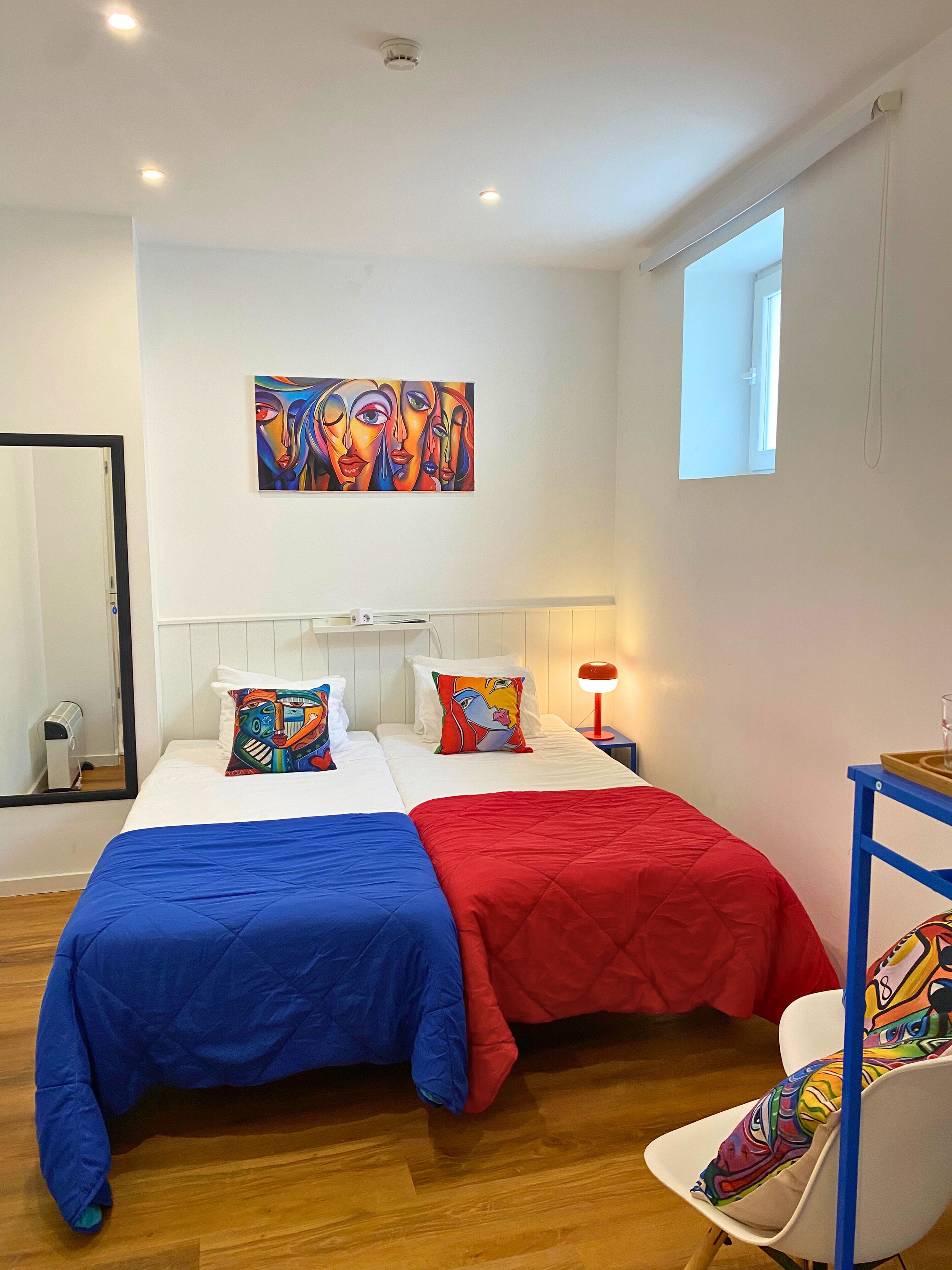 A hotel room with two beds, a colorful painting on the wall, a window, and a chair.