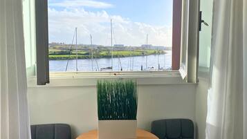 Quarto com vista para um rio e barcos, duas cadeiras cinzentas, uma pequena mesa redonda com um controle remoto e um vaso de plantas.