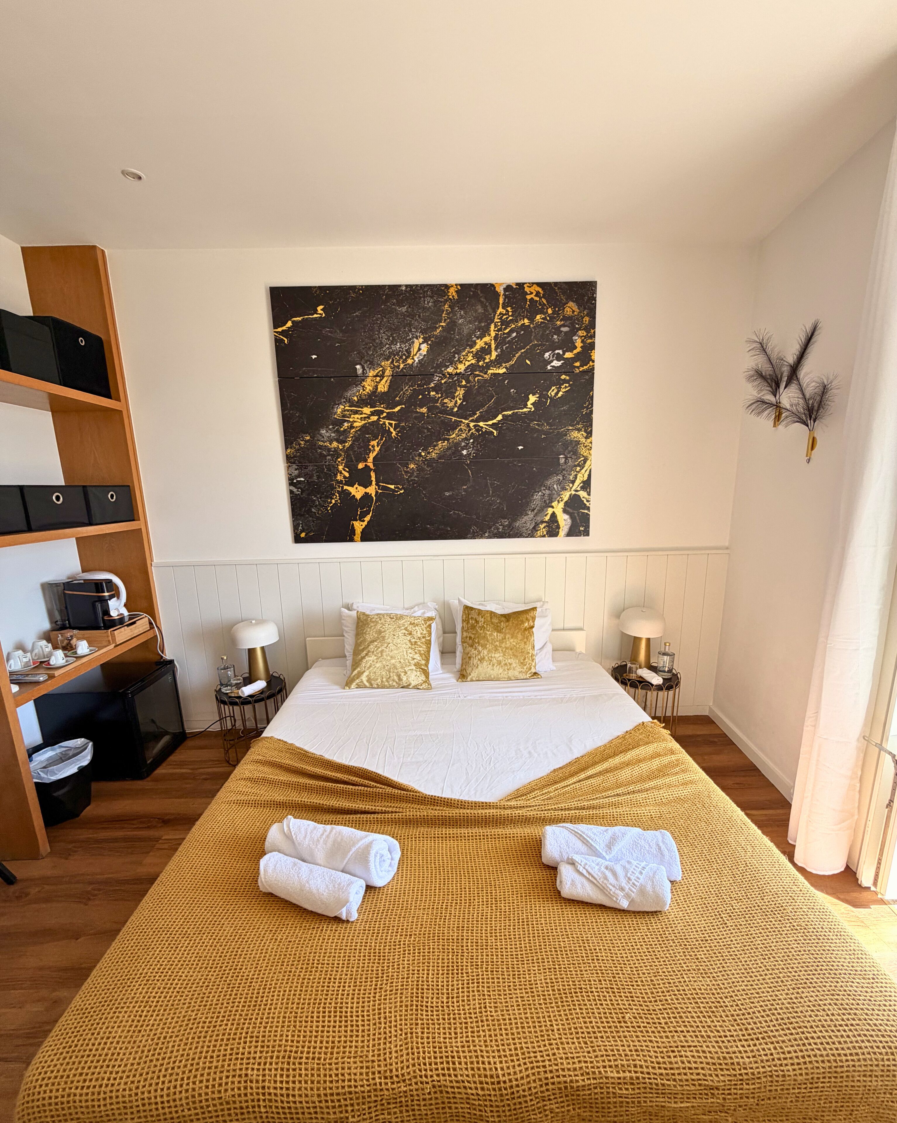 A hotel room with a large bed, a wall-mounted artwork, a bedside table with a lamp, and a shelf with storage boxes.