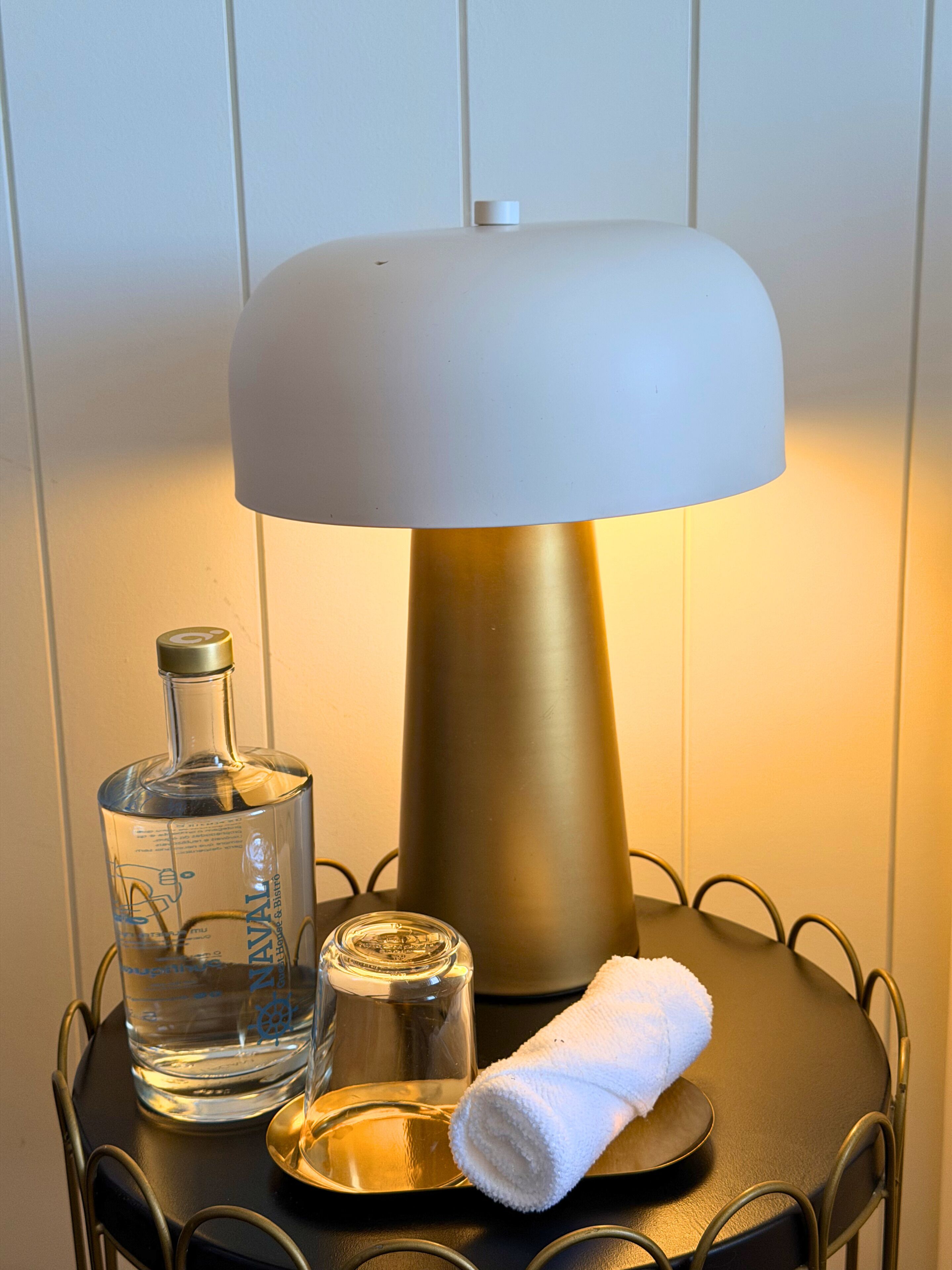 Une lampe avec un abat-jour blanc et un pied doré, une bouteille d’eau, un verre et une serviette enroulée sur une chaise en métal.