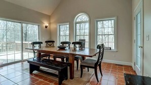 A spacious dining area with a large wooden table, six chairs, and a bench. There are four large windows providing natural light and a view of the outdoors.