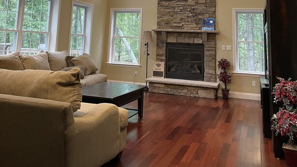 A spacious living room with a stone fireplace, large windows, a ceiling fan, and a cozy seating area.