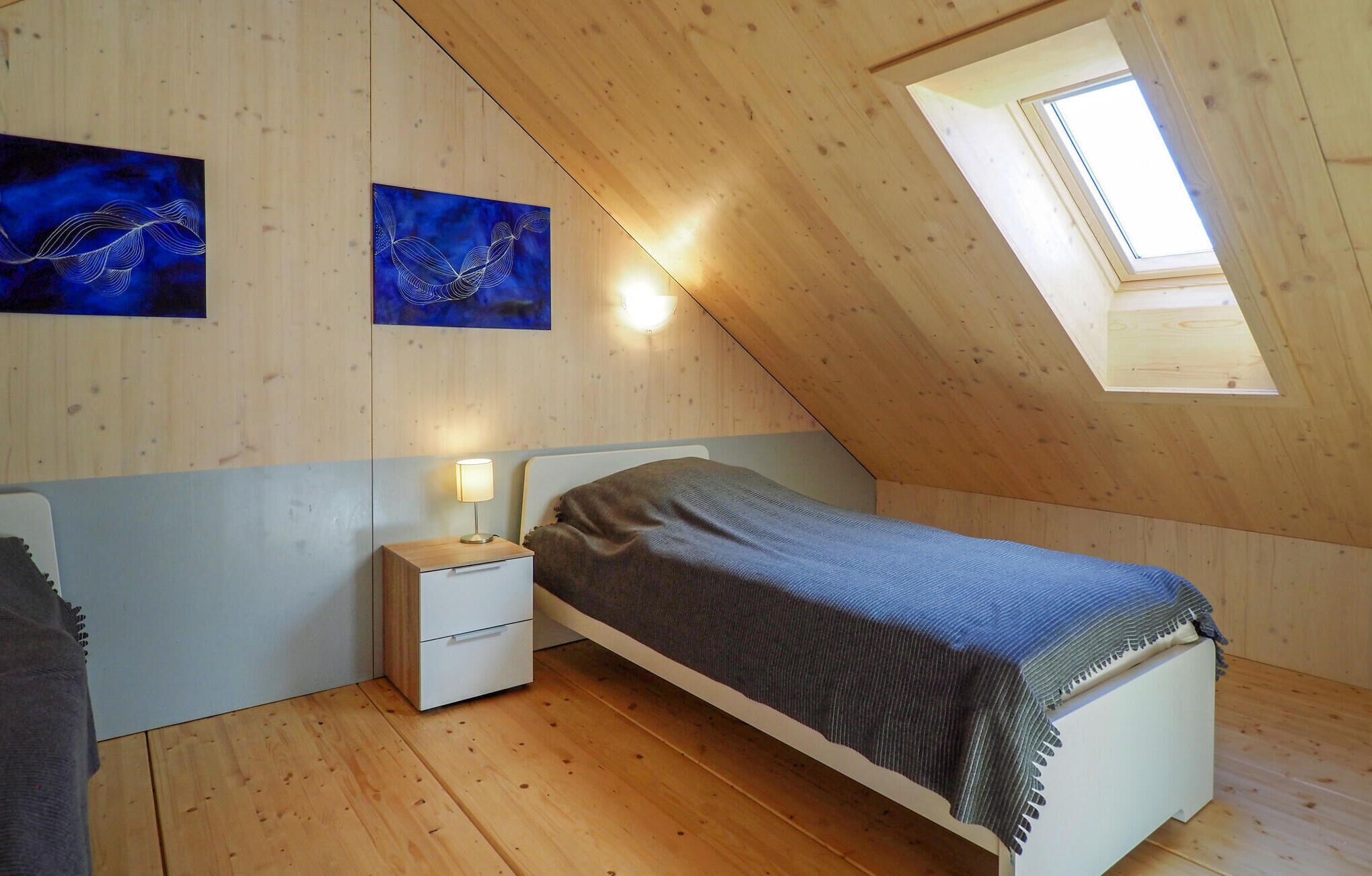 Un dormitorio con techo inclinado, paredes de madera, una cama con manta, una mesita de noche con lámpara y dos cuadros colgados en la pared.
