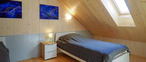 Un dormitorio con techo inclinado, paredes de madera, una cama con manta, una mesita de noche con lámpara y dos cuadros colgados en la pared.