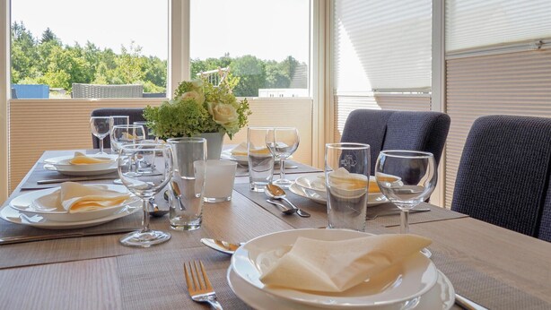 Une table à manger dressée pour deux personnes, avec assiettes, couverts et verres, un vase rempli de fleurs et une vue sur la verdure à travers de grandes fenêtres.