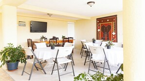 A room with white tables and chairs, a flat-screen TV on the wall, and a decorative wall hanging. - La Maison 29 (Roseau)
