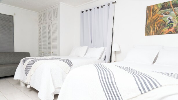 A bedroom with two beds, a grey sofa, white curtains, and a painting on the wall. - La Maison 29 (Roseau)