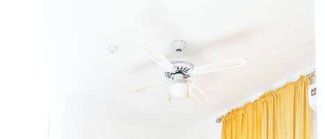 Un dormitorio con una cama de madera, un ventilador de techo, una cortina amarilla y una pared blanca con un adorno.