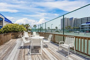 A wooden deck with white metal chairs and a table, overlooking a tennis court and buildings. - White Sands and Good Times (Orange Beach)