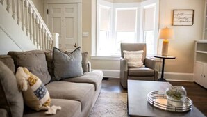Una sala de estar con un sofá color beige, una mesita redonda pequeña y un sillón. Se observa una escalera con barandillas blancas, una puerta y una ventana con vistas.