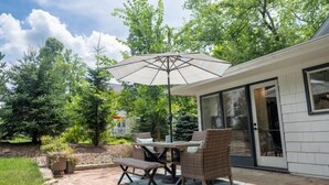 Un patio al aire libre con una mesa, sillas, un paraguas grande y un ventanal corredizo que da a una zona cubierta.