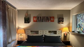 Un dormitorio con una cama, dos mesitas de noche, un ventanal con vistas y una pared decorada con obras de arte.