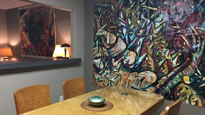 Un comedor con una mesa de madera, cuatro sillas, una lámpara y un cuadro abstracto de colores en la pared.