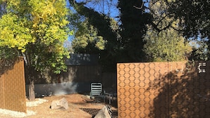 Un espacio al aire libre con suelo empedrado, pared de ladrillo, banco de metal y un árbol.