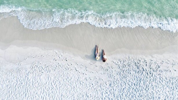 An aerial view of a beach with two people lying on the sand near the water's edge. - Celebrate Mother`s Day on the Beach (Navarre)